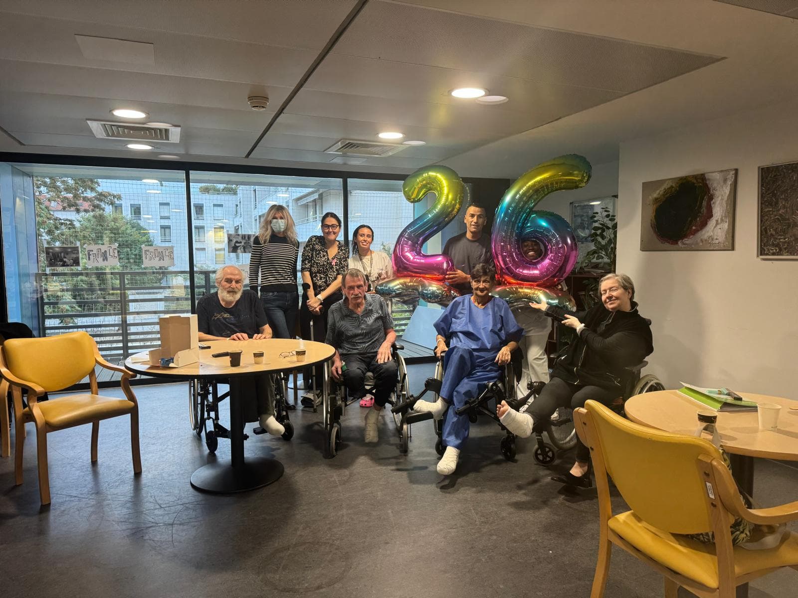 Patients, soignants et visiteurs bénévoles réunis autour de ballons dans la salle de convivialité d'un service hospitalier.