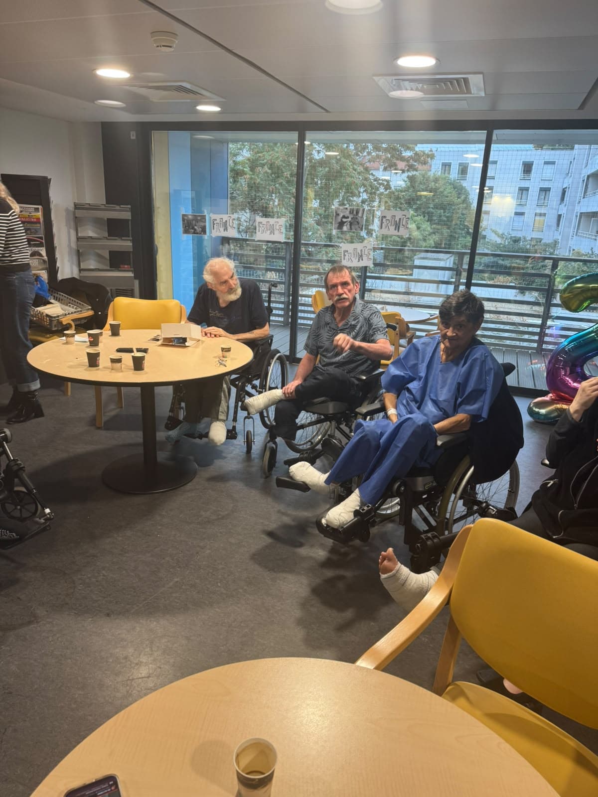 Un visiteur bénévole discute avec des patients installés autour d'une table dans la salle de convivialité.