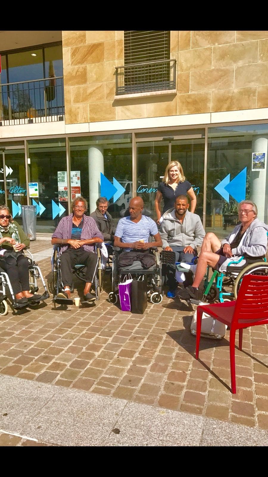 Un groupe de patients en fauteuil roulant et leurs accompagnateurs devant l'entrée vitrée d'un hôpital.